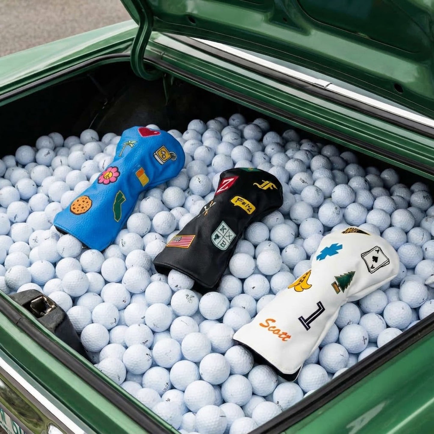 Three custom golf driver head covers in light blue, black, and white in vintage car trunk filled with golf balls - featuring personalized patches including palm tree, hibiscus flower, American flag, name monogram, ace of spades, pine tree, and number 1 - KAMP BANANA handmade head covers