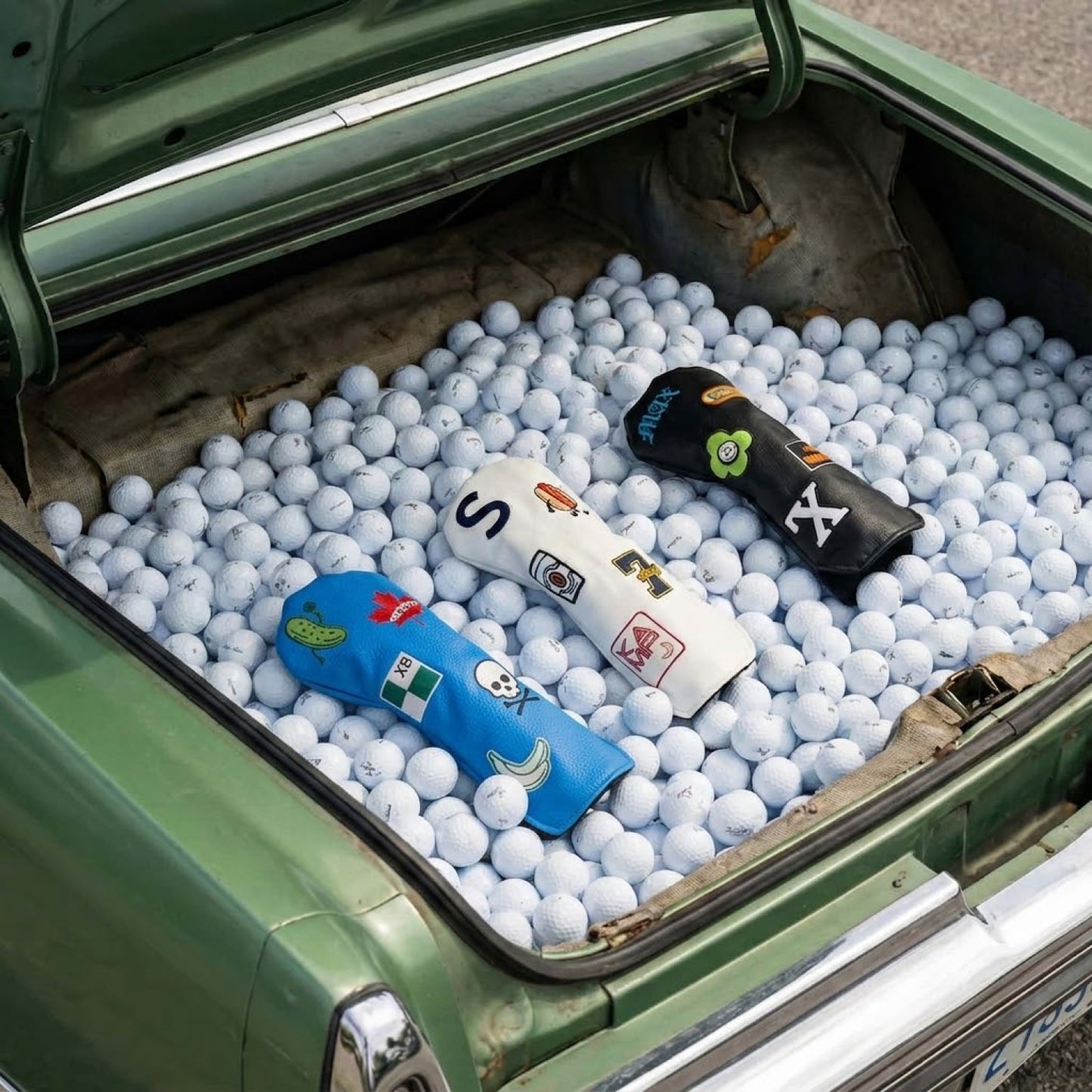 Three custom golf hybrid head covers in light blue, white, and black displayed in vintage car trunk filled with golf balls - featuring personalized patches including Canadian maple leaf, skull and crossbones, pickle, hot dog, and letter monograms - KAMP BANANA handmade head covers