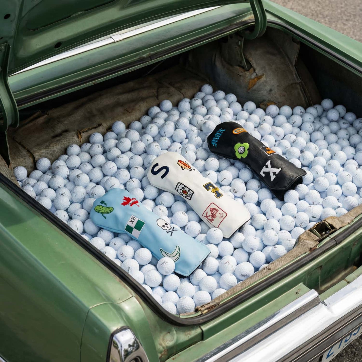 Three custom golf hybrid head covers in light blue, white, and black displayed in vintage car trunk filled with golf balls - featuring personalized patches including Canadian maple leaf, skull and crossbones, pickle, hot dog, and letter monograms - KAMP BANANA handmade head covers