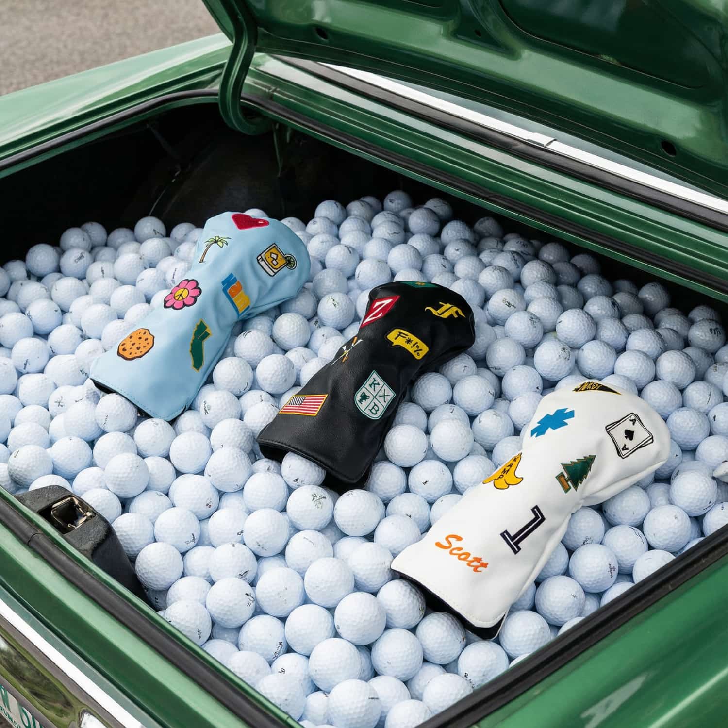 Three custom golf driver head covers in light blue, black, and white in vintage car trunk filled with golf balls - featuring personalized patches including palm tree, hibiscus flower, American flag, name monogram, ace of spades, pine tree, and number 1 - KAMP BANANA handmade head covers