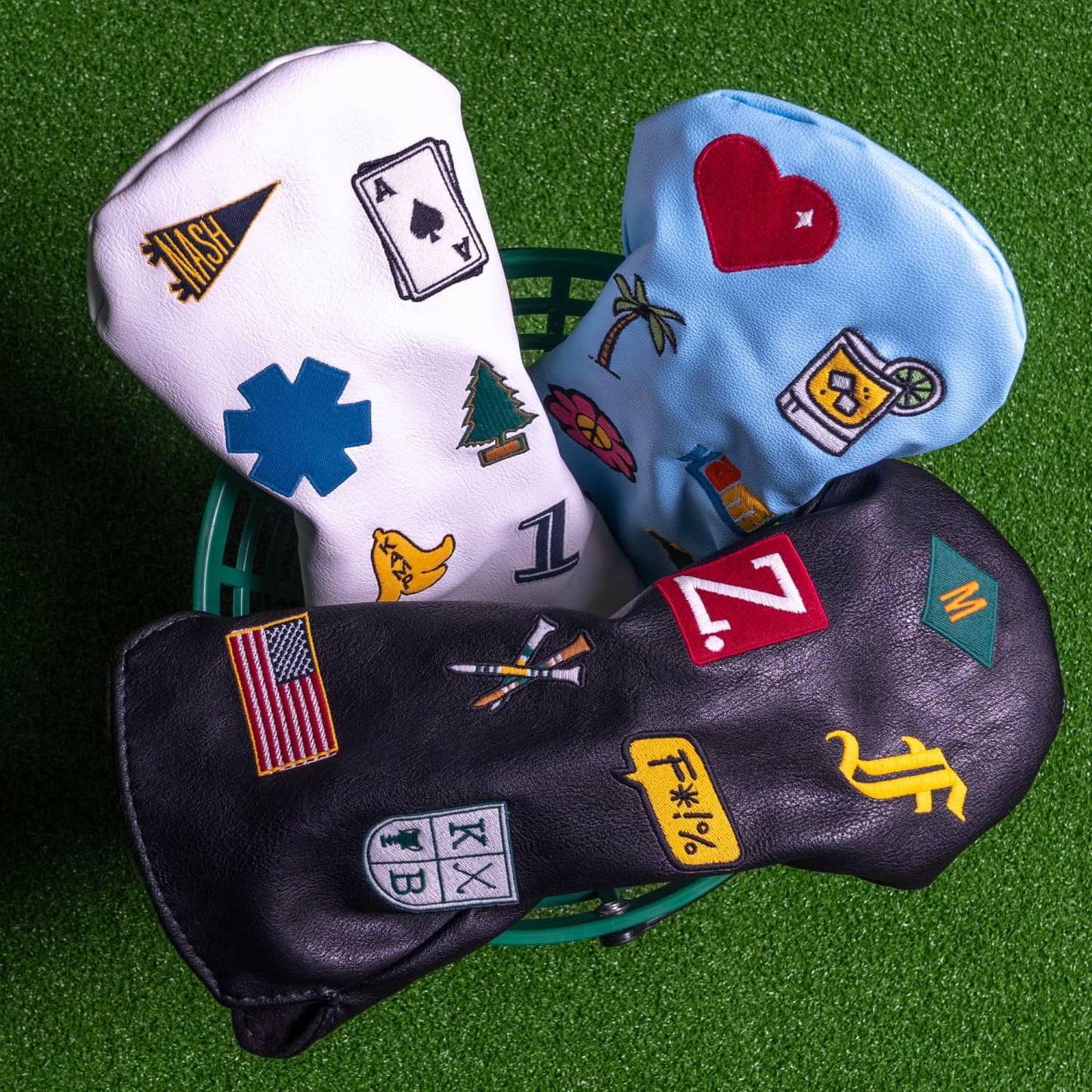 Three custom golf head covers in white, light blue, and black displayed in golf ball basket on green turf - featuring personalized embroidered patches including Nashville pennant, ace of spades, red heart, palm tree, American flag, letter monograms, and pine tree - KAMP BANANA handmade vegan leather head covers