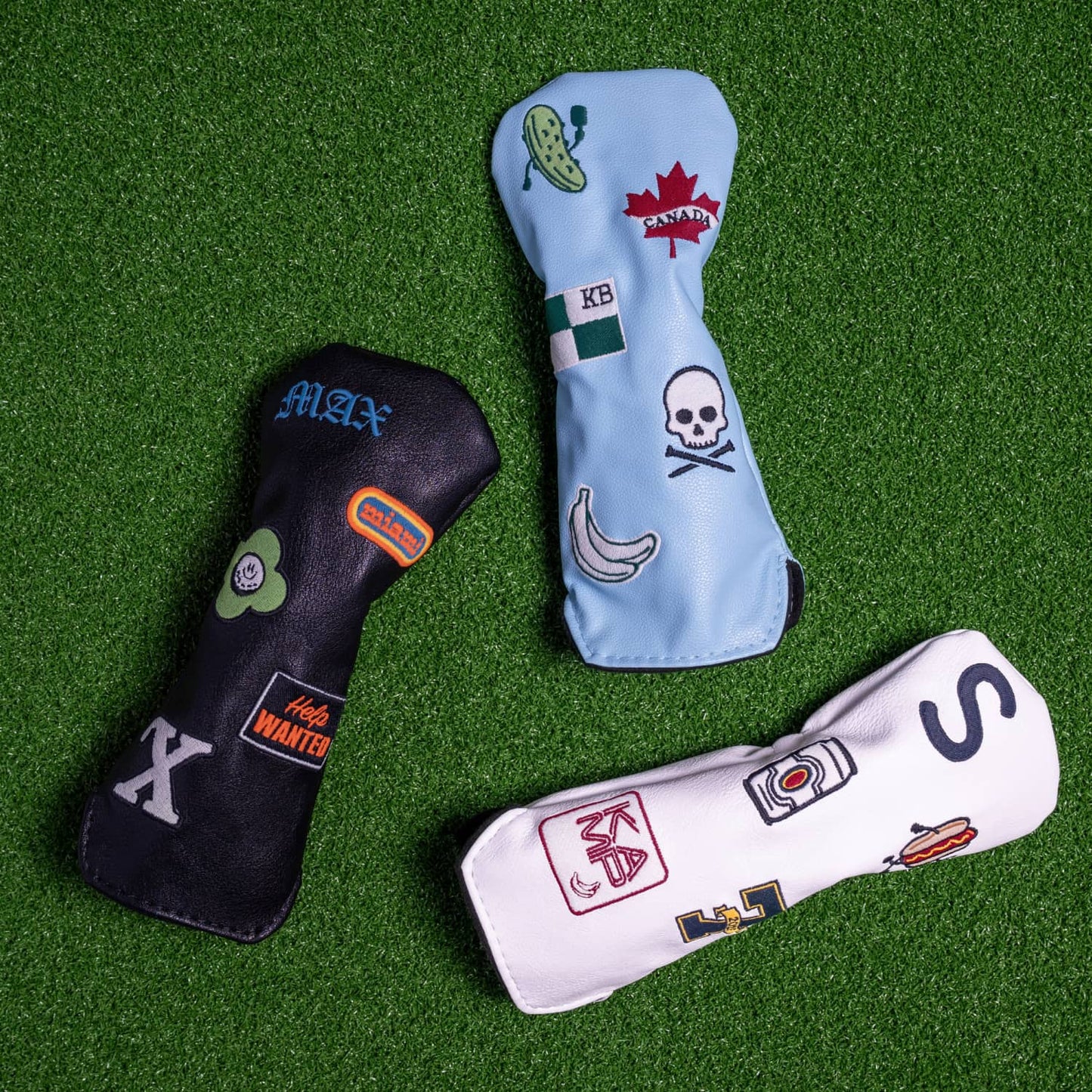 Custom hybrid head covers displayed on green turf - white, light blue, and black vegan leather with personalized patches including monograms, food themes, and travel designs