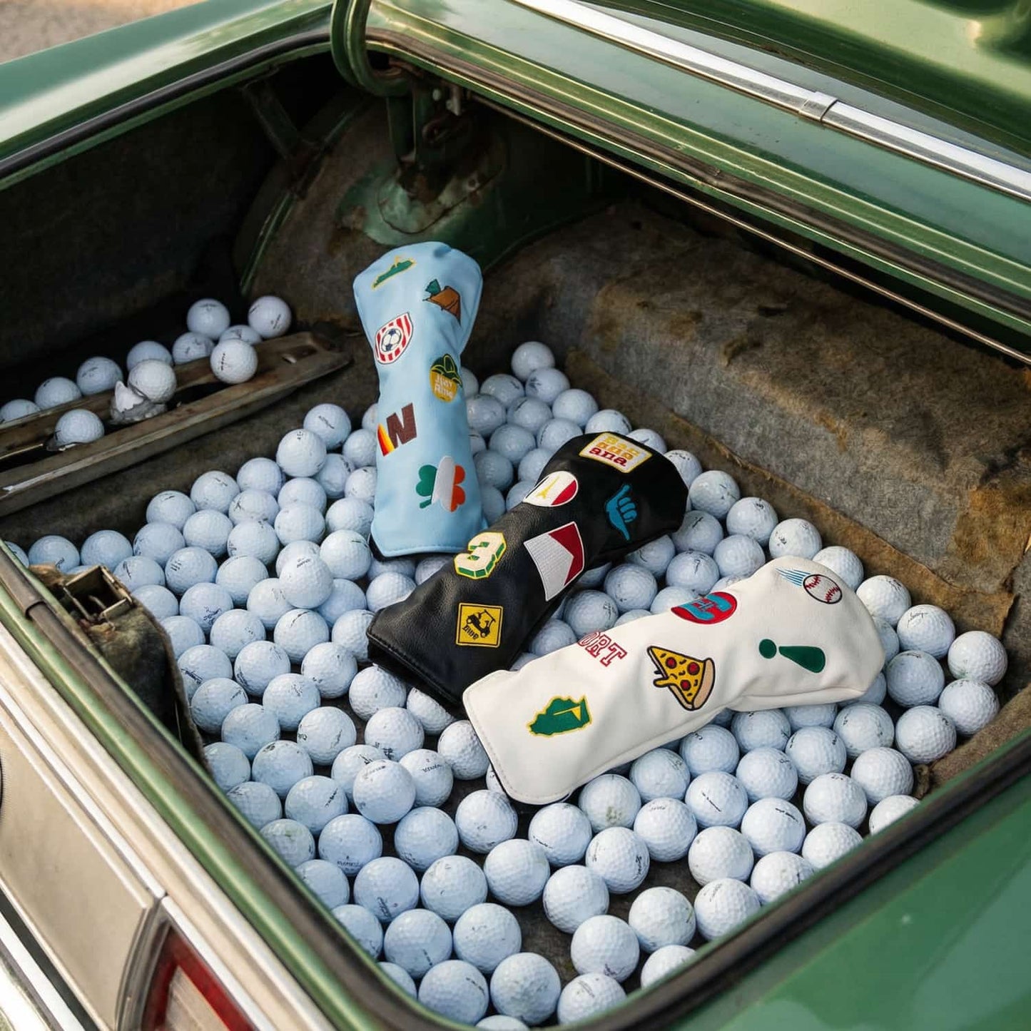 Three custom golf fairway wood head covers in light blue, black, and white displayed in vintage car trunk with golf balls - featuring embroidered patches including Irish shamrock, pizza slice, Kentucky state, camping tent, and golf cart crossing sign - KAMP BANANA personalized head covers