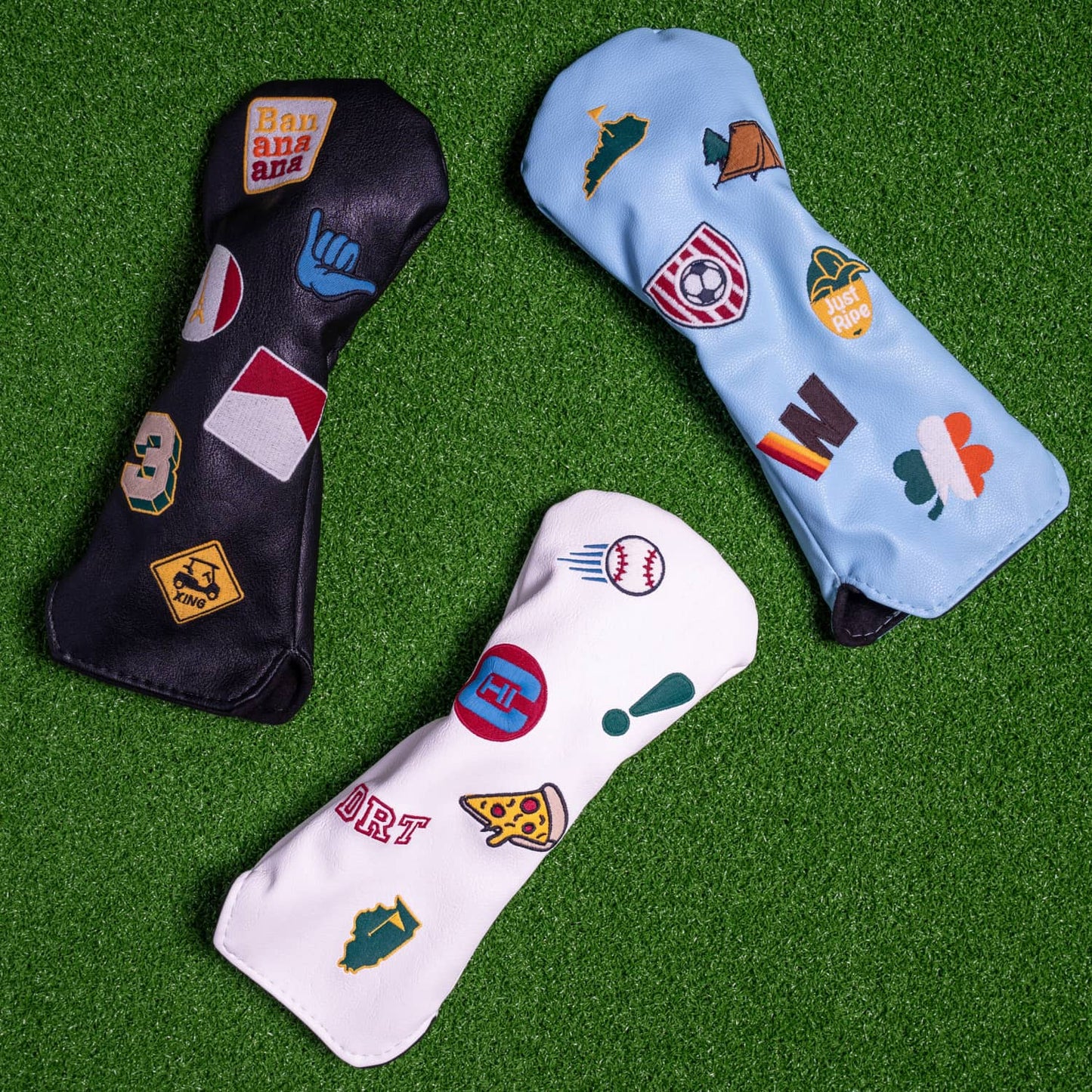 Custom fairway wood head covers displayed on green turf - white, light blue, and black vegan leather with personalized embroidered patches including state flags, sports, and travel themes